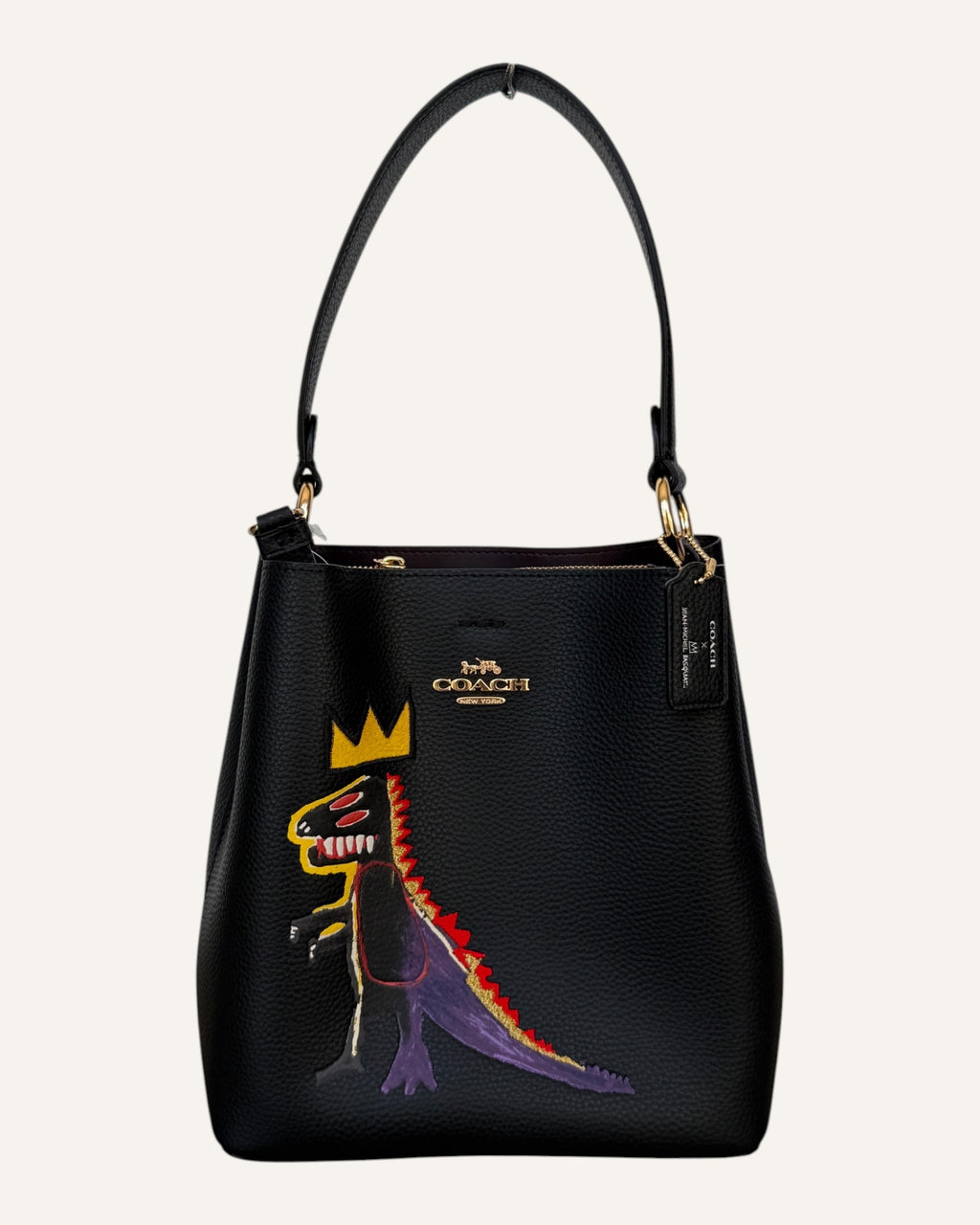 Close-up of a black pebbled leather Coach x Jean-Michel Basquiat bucket bag showing the crowned dinosaur artwork and gold-tone Coach New York logo plaque.