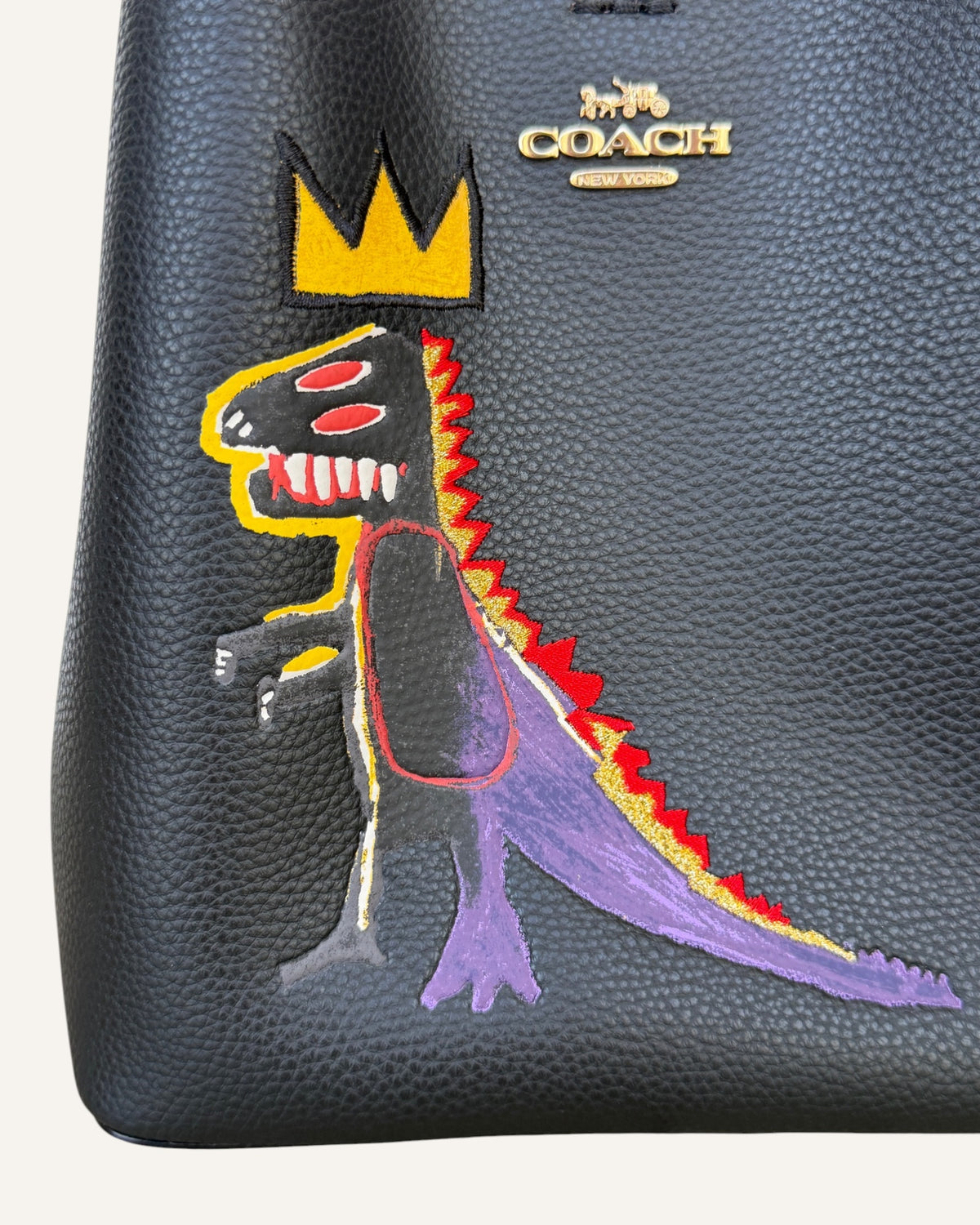 Close-up of a black pebbled leather Coach x Jean-Michel Basquiat bucket bag showing the crowned dinosaur artwork and gold-tone Coach New York logo plaque.