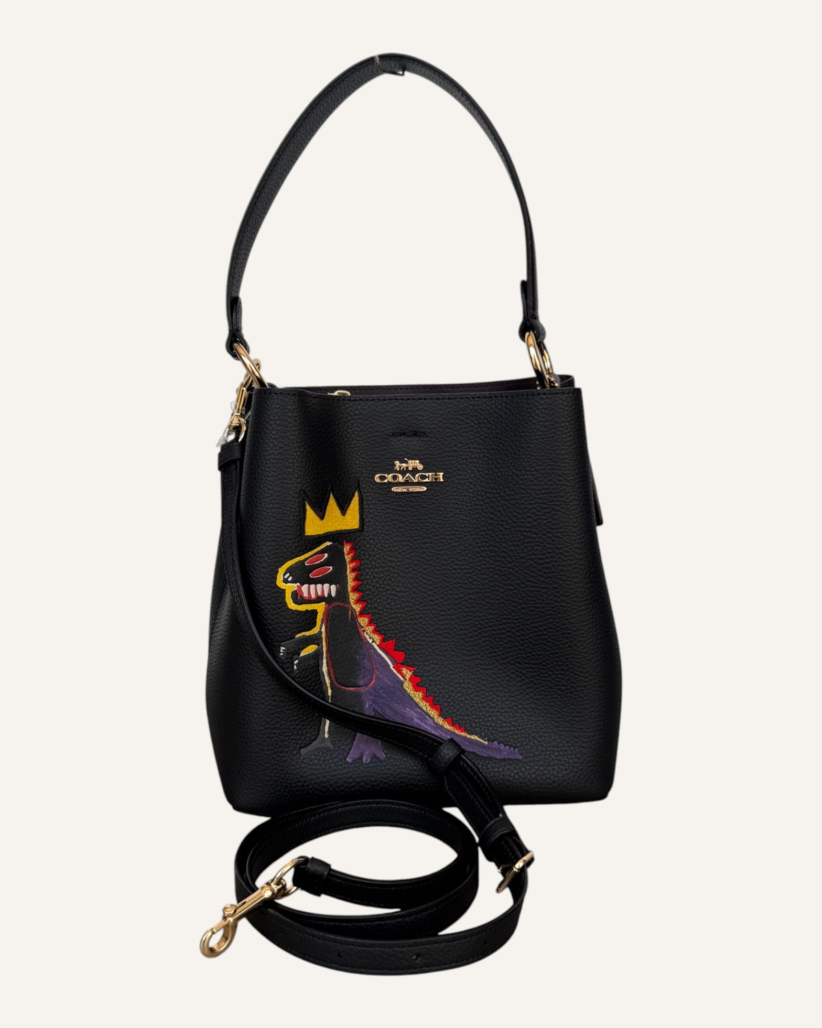 Close-up of a black pebbled leather Coach x Jean-Michel Basquiat bucket bag showing the crowned dinosaur artwork and gold-tone Coach New York logo plaque.