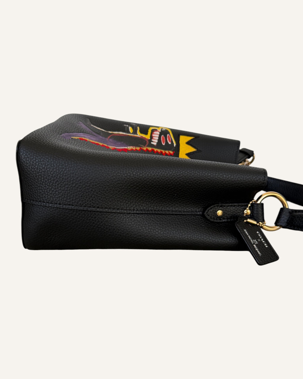 Close-up of a black pebbled leather Coach x Jean-Michel Basquiat bucket bag showing the crowned dinosaur artwork and gold-tone Coach New York logo plaque.